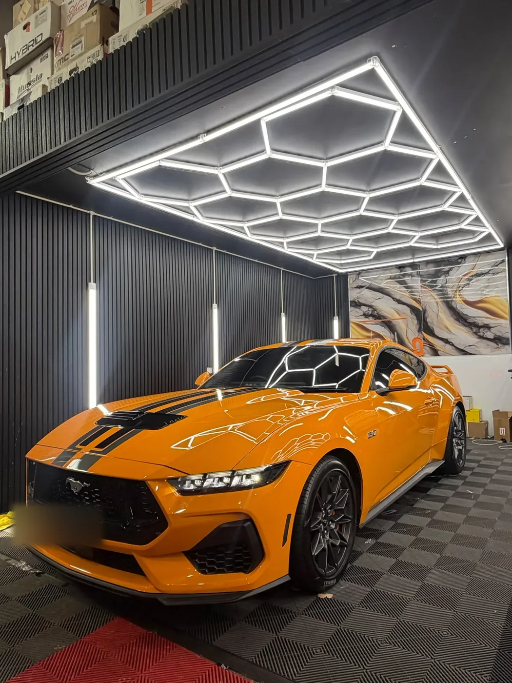 Orange coupe with black stripes inside the studio bay under hexagonal ceiling lights.
